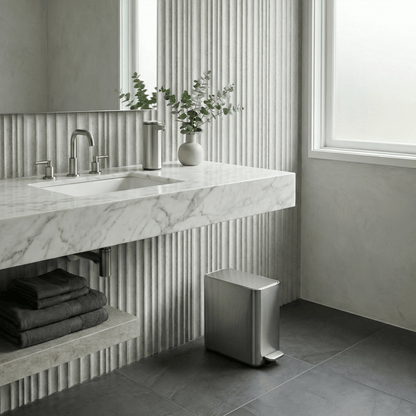 Simplehuman's 9L Slim Step Can in Brushed in a modern, simple bathroom setting. Textured walls with stone flooring and a Simplehuman Sensor Pump in Brushed on the marble sink countertop.