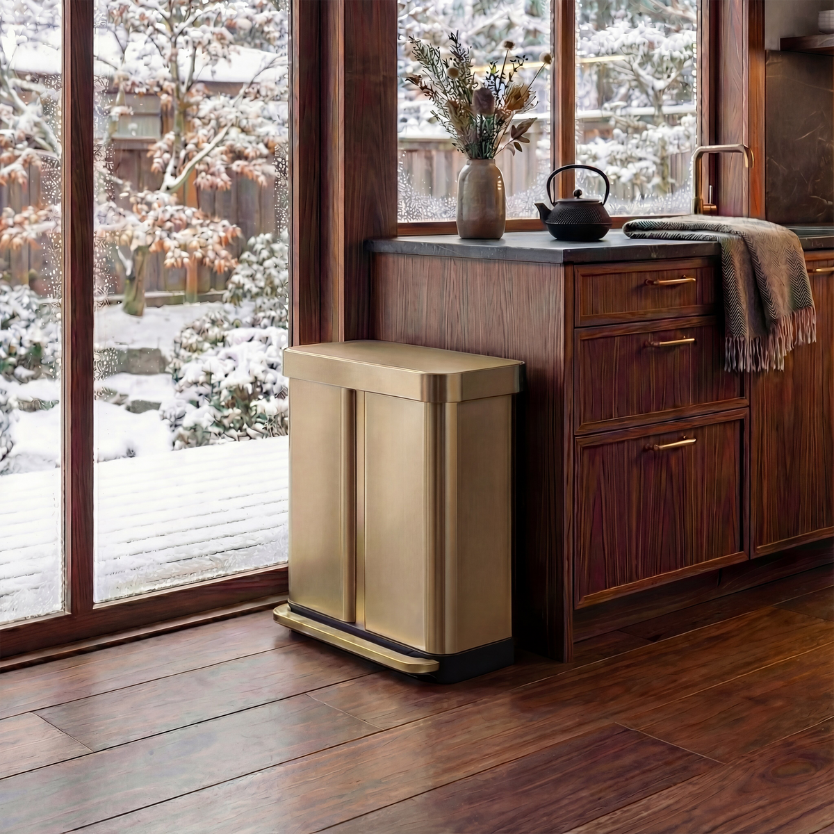 Simplehuman's Matte Gold 58L Step Recycler in a warm, wooden cabin styled kitchen. Surrounded by a tea pot, plants, wooden cabinets, and a view outside with snow on trees.
