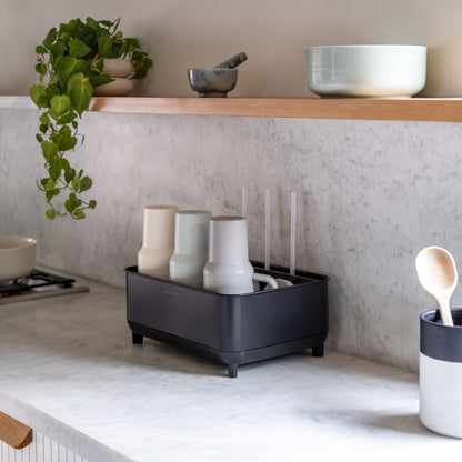 Simplehuman Matte Black Bottle Dock on marble kitchen countertop holding bottles and mugs beside a plant and utensil holder.