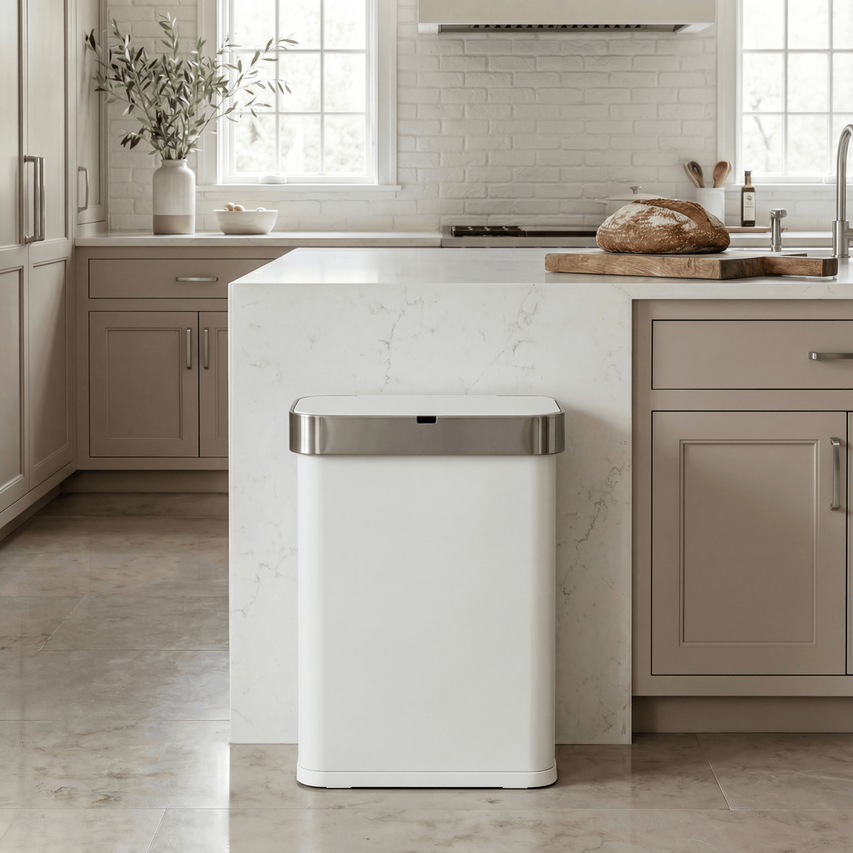 Simplehuman's 58L Sensor Can in White in a modern kitchen setting with neutral cabinetry, white marble counter tops and walls, with slight greenery and natural light in the background.
